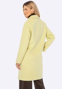 Abrigo de lana amarillo largo con un cuello clásico y una silueta recta. Presenta mangas largas y un mínimo detalle de costuras.