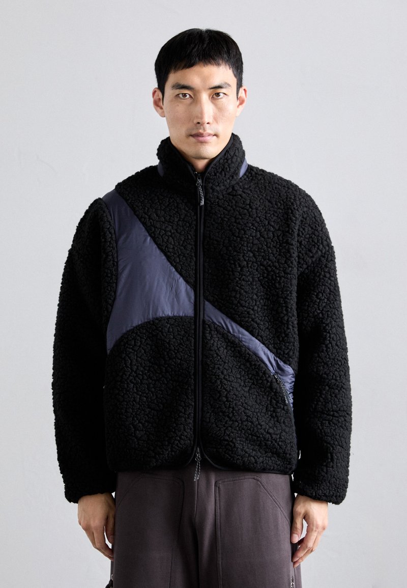 Giacca in fleece nera con finitura testurizzata, caratterizzata da un pannello laterale blu navy liscio. Collo alto, zip a tutta lunghezza e design rilassato.