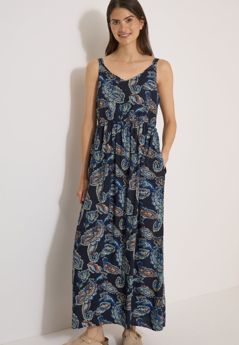 Femme en robe maxi bleu marine sans manches avec motif cachemire multicolore, debout devant un mur clair uni.