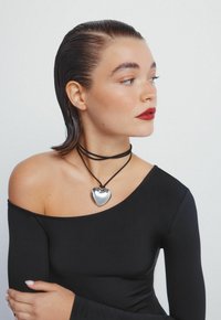 Zwarte long sleeve top, zilveren hart hangerketting aan een zwarte koord, gladde textuur, model gepresenteerd in zijprofiel.