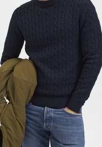 Marinblå kabelstickad tröja med rund halsringning, ribbstickade ärmslut och nederkant, tillsammans med en olivgrön jacka som hänger över ena armen.