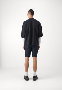 Svart oversized t-shirt med korta ärmar, som är ihopkopplad med marinblå shorts. Modellen bär vita strumpor och svarta skor, stående mot en enkel bakgrund.