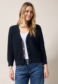 Námořnický pletený cardigan s výstřihem do V, šesti velkými knoflíky a ribovanými manžetami a lemem. Spárováno s bílým tričkem a modrými džíny.