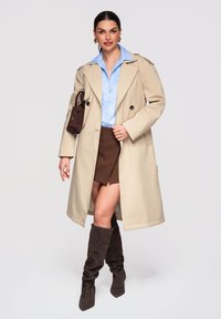 Femme portant un trench-coat beige sur une chemise bleue et une mini-jupe marron, tenant un petit sac à main marron et portant des bottes marron souples montant jusqu'aux genoux.