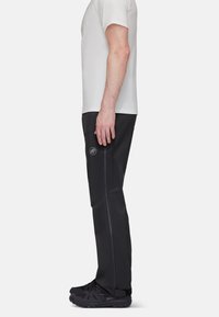 Svarta utomhusbyxor med matt finish, med fickor som stängs med dragkedja och en logotyp på låret. Bärs med en ljusgrå t-shirt och svarta skor.