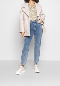 Beige kabát ze směsi vlny s širokými klopami, zelená květinová blůza, modré džíny s vysokým pasem a šedo-bílé tenisky. Modelka pózuje čelně.