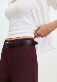 Pantaloni tailleur bordeaux con tasche laterali e una cintura in pelle nera. La camicia è bianca, con bottoni, e indossata in modo leggero sopra.