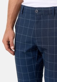 Marineblauwe geruite broek met een subtiele geweven textuur. Voorzien van een enkele knoopsluiting en zijzakken. Het patroon bestaat uit witte rasterlijnen.