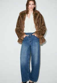 Abrigo de piel sintética marrón sobre una blusa blanca, combinado con jeans azules de pierna ancha y talle alto. Calzado casual, diseño sencillo, colores neutros.