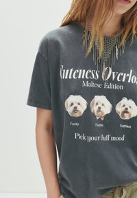 T-shirt en coton gris présentant trois visages de chiens maltais avec le texte : "Surcharge de mignonnerie", "Choisissez votre humeur en peluche." Comprend un collier en chaîne superposé.