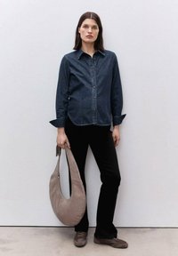 Jeansskjorta med knappar, mörkblå; svarta byxor; beige hobo-väska; bruna skor; modell står mot en neutral bakgrund, armarna avslappnade.