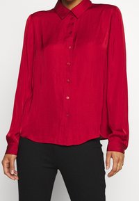 Červená saténová blůza s límečkem, předním zapínáním na knoflíky a dlouhými rukávy. Má měkkou strukturu a mírně zakřivený lem.
