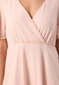 Rochie roz pal cu decolteu în V, mâneci scurte și talie strânsă, realizată dintr-un material moale, fluid, cu o textură netedă.