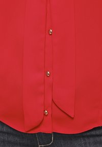 Röd blus i ett mjukt, lätt tyg med guldknappar och en framknäppning. Lätt rundad fåll och mjuk textur.