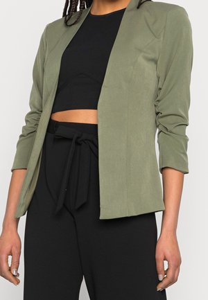 Personne portant un blazer vert olive à manches froncées par-dessus un crop top noir et un pantalon noir taille haute noué avec une ceinture en tissu.