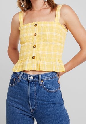 Kobieta w żółtym kraciastym, bezrękawnym marszczonym topie z guzikami i niebieskich wysokich dżinsach, stojąca z rękami za plecami.