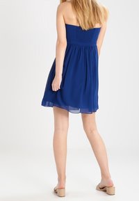 Modrá bezramínková šaty s smocked tělem a volnou sukní. Lehká tkanina; délka do poloviny stehna. Nosené s nahými sandály.