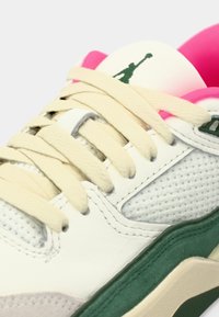 Detailní záběr na bílé tenisky s béžovými tkaničkami, zelenou podrážkou, růžovou vnitřní výstelkou a zeleným logem Jumpman na jazyku.