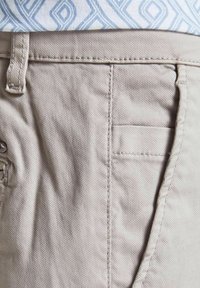 Světlé béžové bavlněné kalhoty s hladkým, texturovaným povrchem, viditelným šitím a boční kapsou. Pozadí zobrazuje vzorovanou tkaninu.