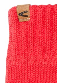Rote Strickmütze mit strukturiertem Muster und einem Holzetikett mit Logo. Das Material wirkt weich und elastisch.