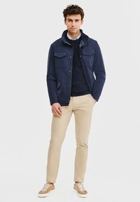 Granatowa kurtka z dwoma kieszeniami na piersi, z zamkiem błyskawicznym i zapięciem na napy, nałożona na granatowy sweter. Beżowe spodnie i brązowe sneakersy dopełniają całość.