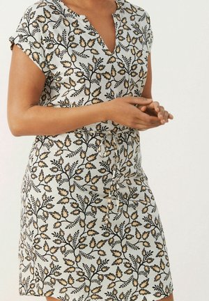 Frau trägt ein kurzärmeliges, V-Ausschnitt-Kleid mit beigem und schwarzem Blumenmuster und Taillenschleife, die Hände vor dem Körper verschränkt vor schlichtem Hintergrund.