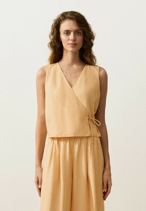 Tânără cu păr creț purtând un top bej fără mâneci în stil wrap și pantaloni largi asortați, stând în fața unui fundal simplu și luminos.