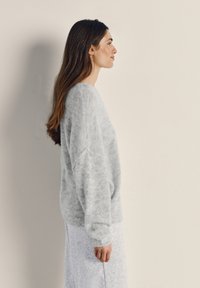 Žena s dlhými hnedými vlasmi, oblečená v svetlo šedom huňatom svetri a zodpovedajúcej sukni, stojaca bokom pri hladkej béžovej stene.