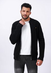 Gilet en tricot noir avec un design texturé, fermeture éclair et un subtil accent doré. Assorti à un t-shirt blanc et un jean sombre.