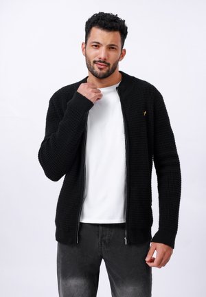 Svart strikket cardigan med teksturert design, glidelås og en subtil gyllen aksent. Kombinert med en hvit t-skjorte og mørke jeans.