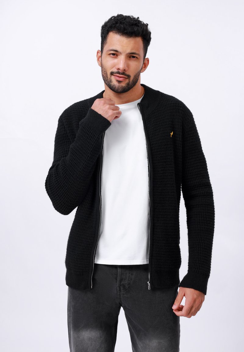 Gilet en tricot noir avec un design texturé, fermeture éclair et un subtil accent doré. Assorti à un t-shirt blanc et un jean sombre.