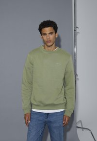 Giovane con capelli ricci che indossa una felpa verde chiaro e jeans blu, in piedi contro uno sfondo grigio da studio.