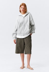 Grå hettejakke med oversized passform, frontlommer, brukt over en hvit skjorte, kombinert med olivenfargede shorts og hvite flip-flops.