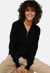 Haut de sweat à capuche noir zippé avec une texture douce et des manches longues, doté d'une capuche et de détails en fermeture éclair argentée, porté avec un pantalon beige clair.