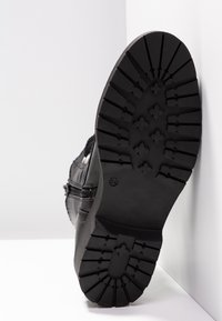 Semelle de botte noire vue de face avec des crampons profonds et des motifs en forme d'étoile, positionnée contre un fond blanc d'angle.