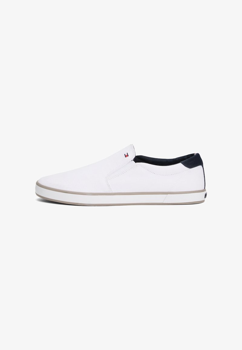 Baskets slip-on blanches avec semelle marron clair, col de talon noir et petit logo rouge, blanc et bleu près de l'ouverture sur fond blanc.