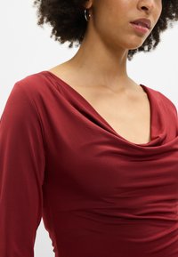 Burgundijska dolgoročna majica s padajočim izrezom, ki se ponaša z mehkim, gladkim materialom in nabranimi detajli na pasu za prilegajoč silhueto.