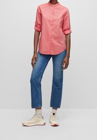 Person iført en rullet ærmes koralfarvet skjorte med knapper, blå jeans, beige sneakers med røde såler og lyse sokker, der står på et gråt gulv.