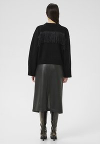 Frau, die einen schwarzen Strickpullover mit Fransen auf dem Rücken, einen perforierten schwarzen Midi-Rock und schwarze Stiefel mit Absatz trägt, steht mit dem Rücken zur Kamera.