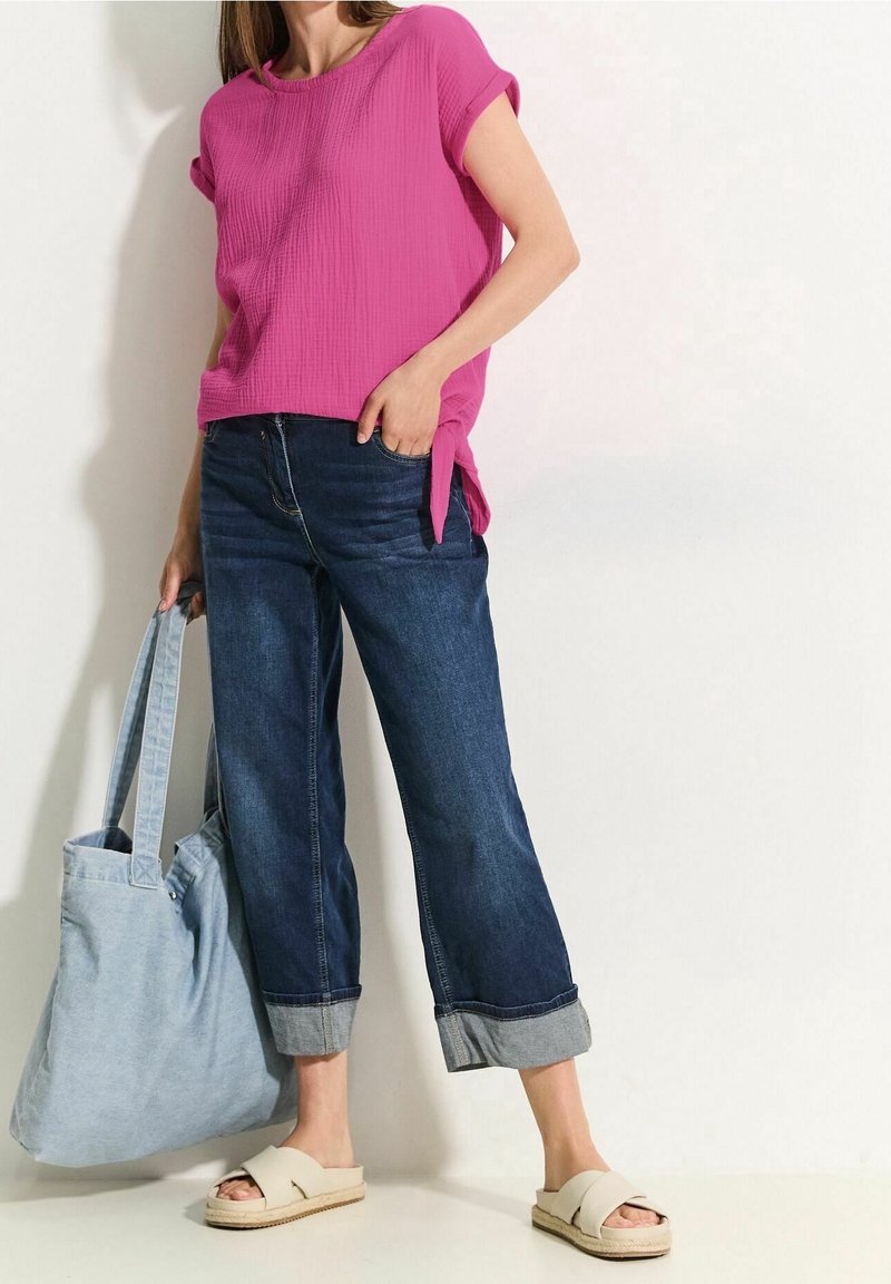 Donna con una maglietta rosa a maniche corte, jeans blu scuro arrotolati, sandali beige e una grande borsa tote azzurro chiaro.