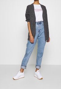 Cardigan long gris, T-shirt blanc avec texte rouge "FEMME", jeans taille haute bleu clair, et baskets blanches avec semelles marron.