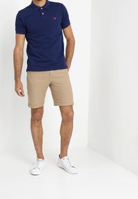 Marinblå pikétröja med knapplist med tre knappar och liten logotyp, kombinerad med beige shorts och vita sneakers. Blandning av bomull, avslappnad passform.