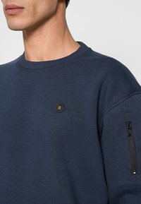 Donkerblauwe sweatshirt met ronde halslijn, geribbelde manchetten en een ritssluiting aan de mouwzak met een hexagonaal logo in goud en zwart.