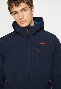 Mørkeblå jakke med hætte, som har orange lynlåse og et logo. Includes to forlommer og et struktureret stof.