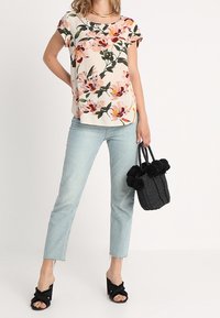 Donna che indossa una blusa a maniche corte floreale, jeans azzurri chiari, sandali neri con tacco, mentre tiene una borsa a tracolla nera intrecciata con pompon.
