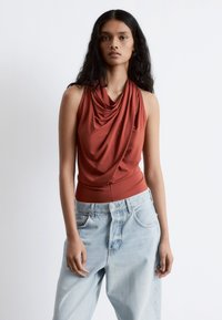 Rostfarbener ärmelloser Oberteil mit drapiertem Ausschnitt, kombiniert mit hellblauen, weiten Jeans. Glattes Material, lockere Passform und lässiger Stil.