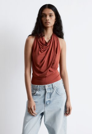 Rust-colored sleeveless top with draped neckline, paired with light blue baggy jeans. Smooth fabric, relaxed fit and casual style.