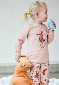 Ungt barn med blont hår i rosa djurtemade pyjamaser dricker ur en juicebox medan det håller en stor brun plyschhäst.