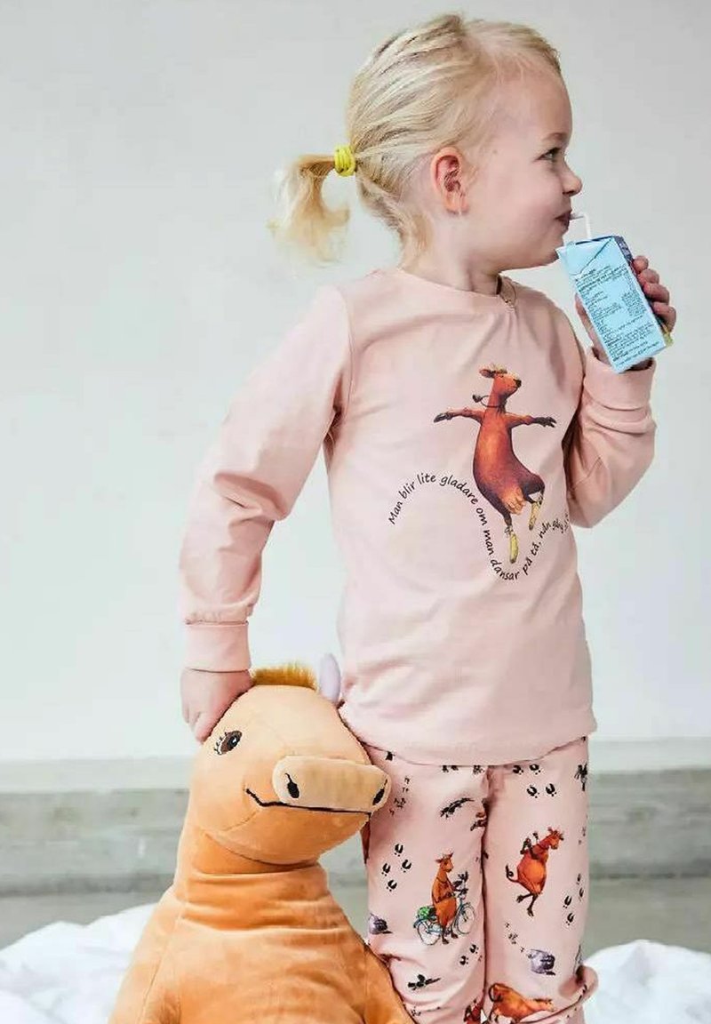 Ungt barn med blont hår i rosa djurtemade pyjamaser dricker ur en juicebox medan det håller en stor brun plyschhäst.
