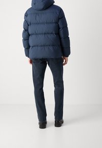 Marinblå puffjacka med horisontellt quiltat mönster, huva och elastiska manschetter, bärs över mörka jeans och svarta skor, visar en bakvy.